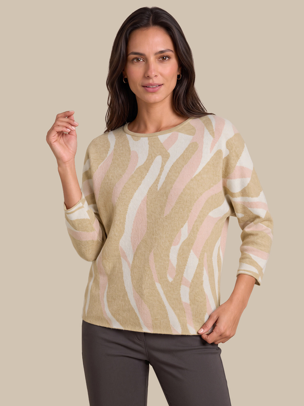 A woman modeling a beige, white, and light pink zebra-patterned knit sweater with three-quarter sleeves, paired with dark gray pants, against a solid beige background.