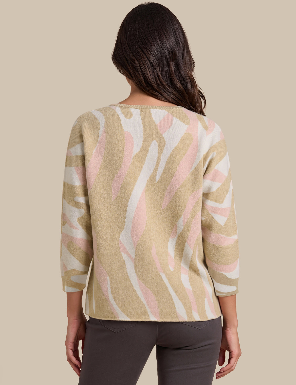 A woman viewed from the back wearing a beige knit sweater with a pink and white abstract wavy pattern, paired with dark gray pants, against a solid beige background.