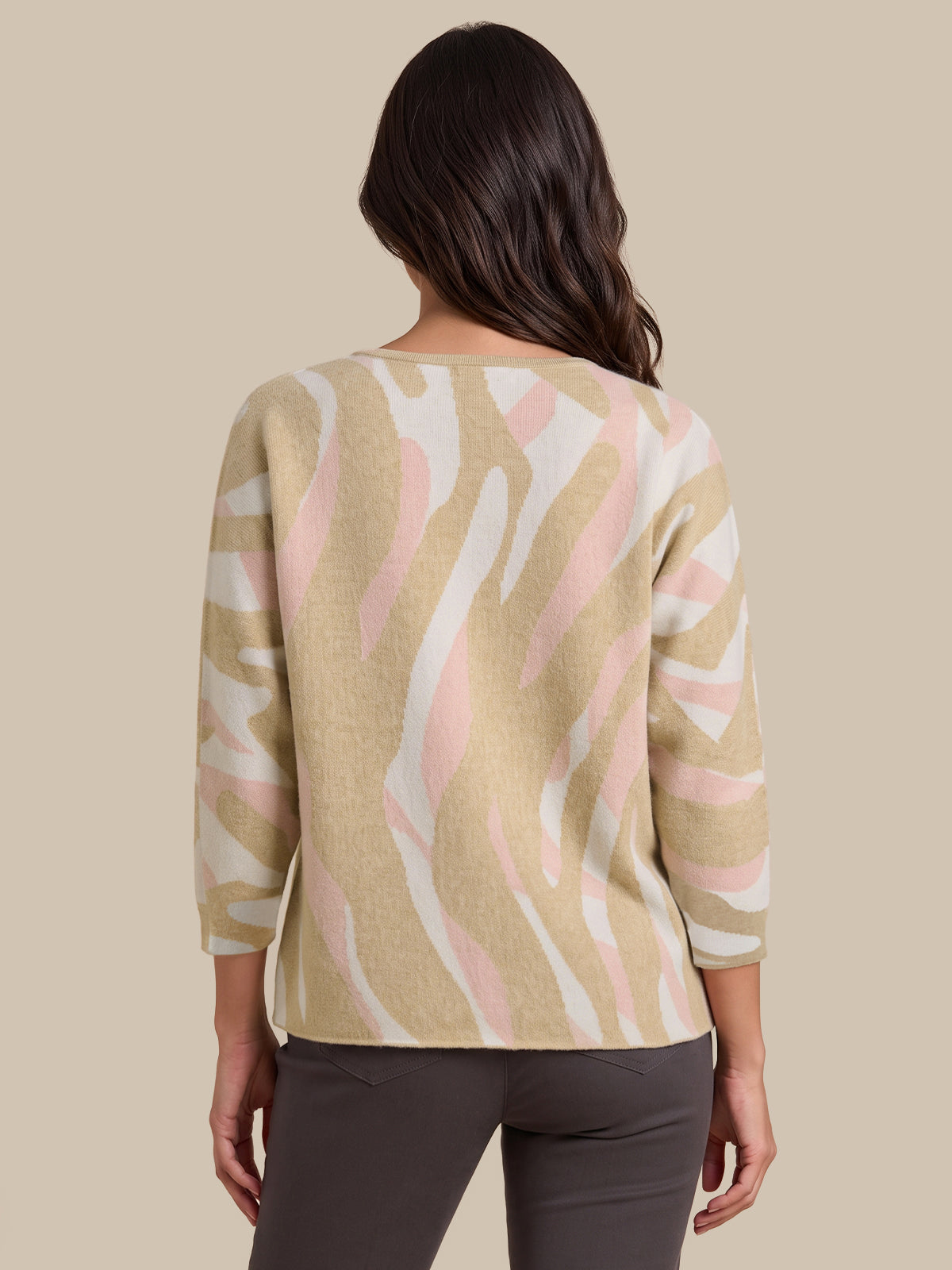 A woman viewed from the back wearing a beige knit sweater with a pink and white abstract wavy pattern, paired with dark gray pants, against a solid beige background.