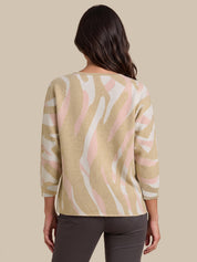 A woman viewed from the back wearing a beige knit sweater with a pink and white abstract wavy pattern, paired with dark gray pants, against a solid beige background.