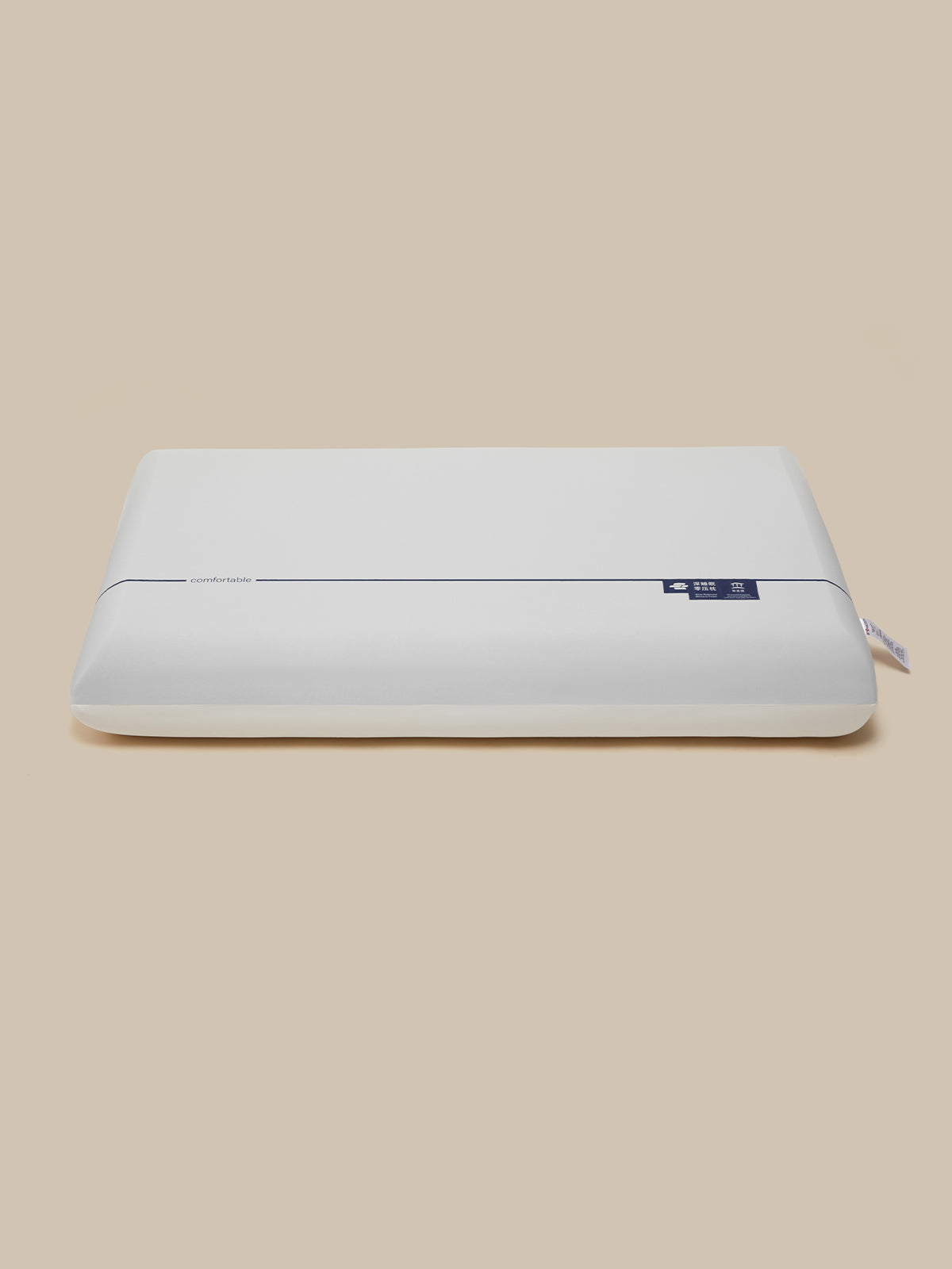 A white rectangular pillow with a smooth, matte finish and a small blue brand tag on the upper right side, set against a solid beige background.