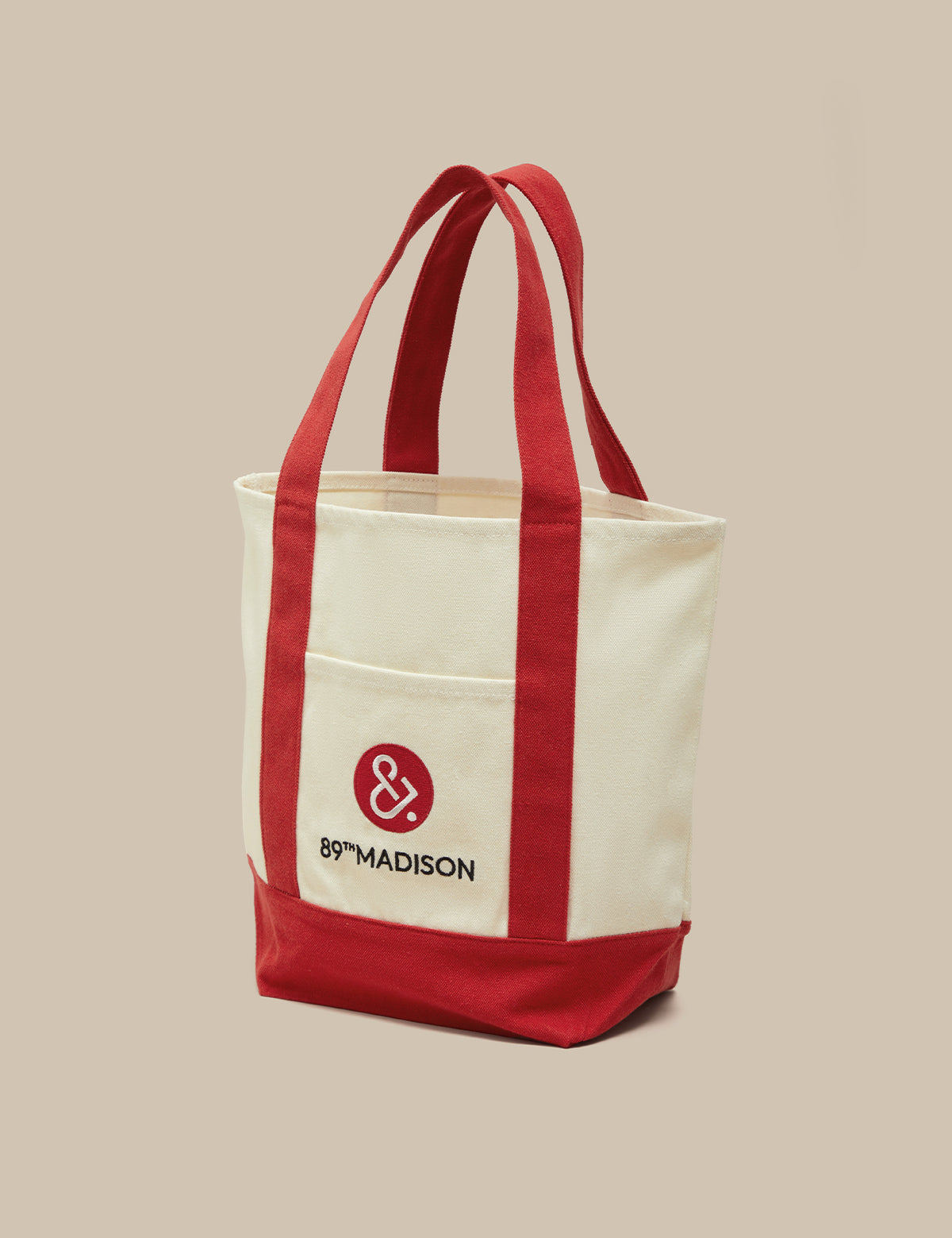 An off-white canvas tote bag with red handles, a red bottom panel, and a red front pocket featuring the '89°MADISON' logo, set against a solid beige background.