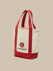 An off-white canvas tote bag with red handles, a red bottom panel, and a red front pocket featuring the '89°MADISON' logo, set against a solid beige background.