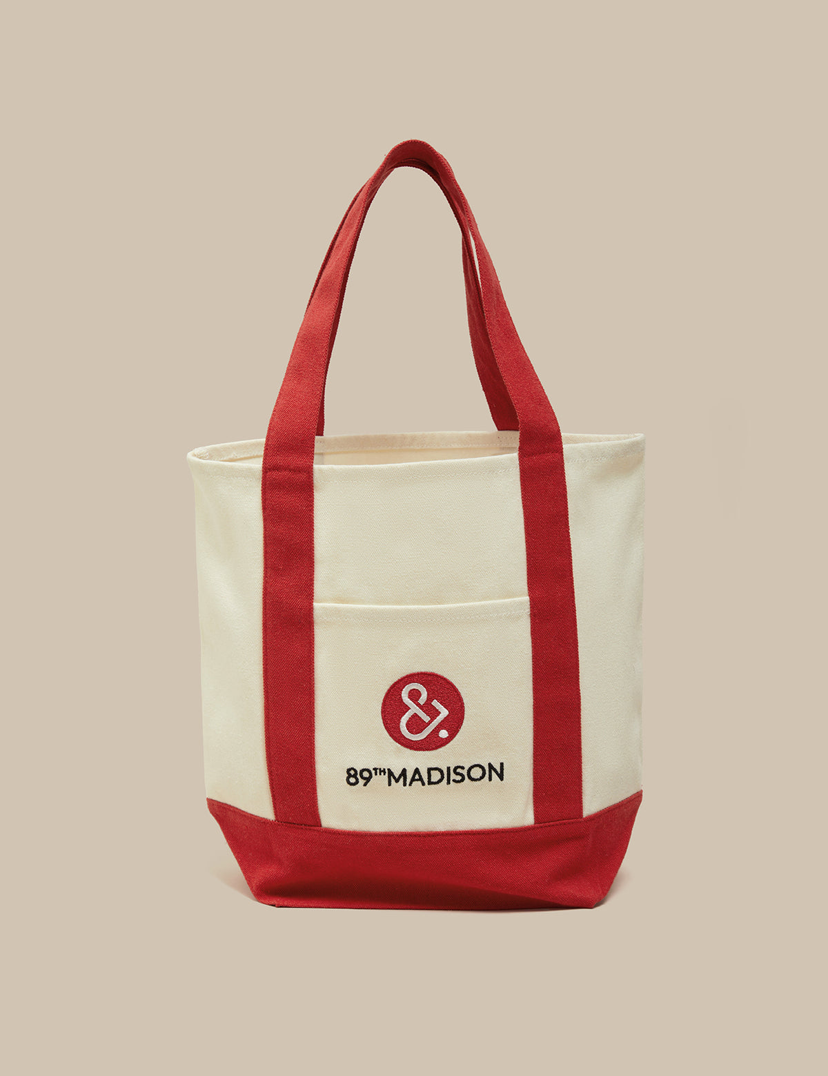 An off-white canvas tote bag with red handles, red side panels, and a red bottom base. The front features a pocket with a red circular logo containing an ampersand and the text '89°MADISON' in black, set against a solid beige background.