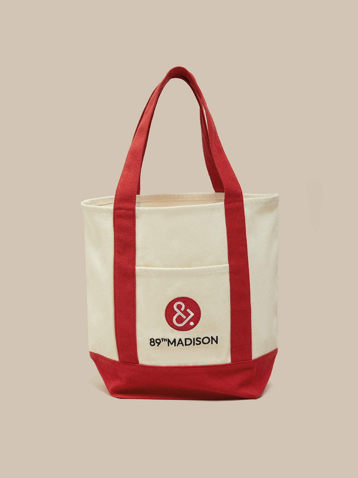 An off-white canvas tote bag with red handles, red side panels, and a red bottom base. The front features a pocket with a red circular logo containing an ampersand and the text '89°MADISON' in black, set against a solid beige background.