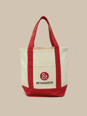 An off-white canvas tote bag with red handles, red side panels, and a red bottom base. The front features a pocket with a red circular logo containing an ampersand and the text '89°MADISON' in black, set against a solid beige background.