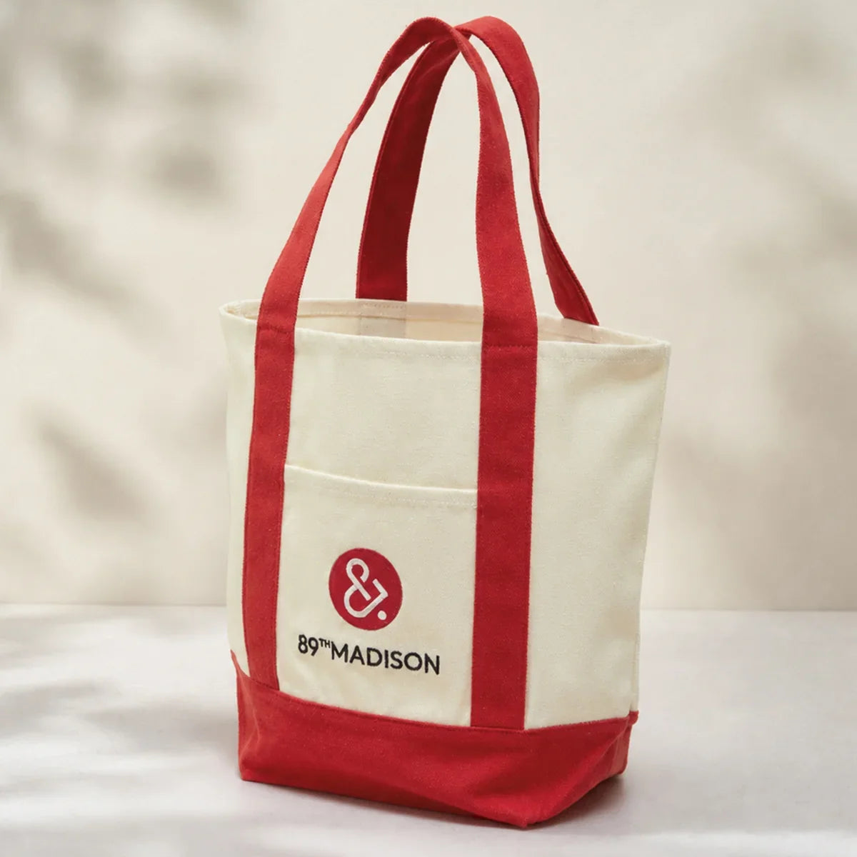 A canvas tote bag with cream-colored body and red handles, bottom trim, and front pocket. The front pocket features a red circular logo with an ampersand and the text '89TH MADISON'.