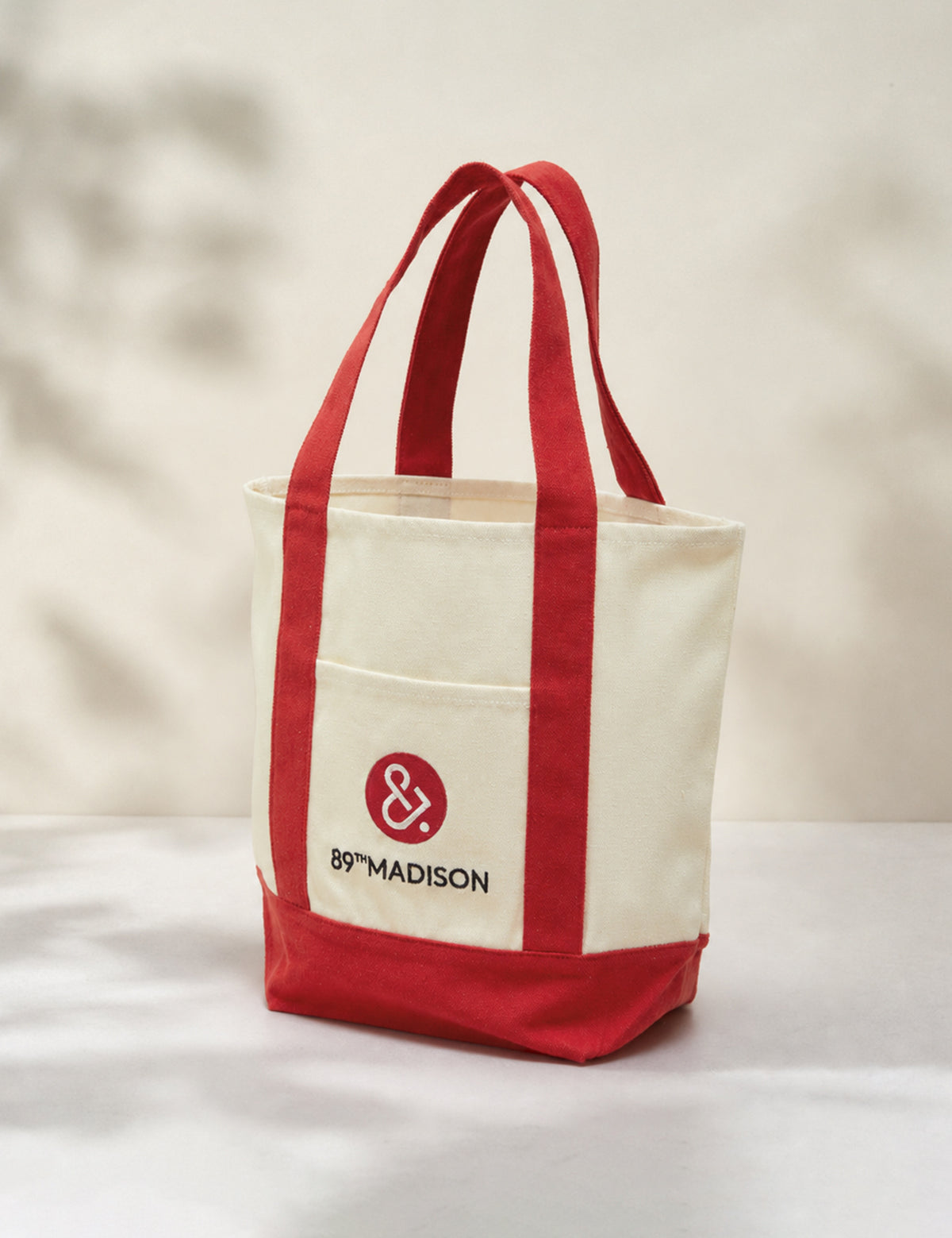 A canvas tote bag with cream-colored body and red handles, bottom trim, and front pocket. The front pocket features a red circular logo with an ampersand and the text '89TH MADISON'.