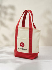 A canvas tote bag with cream-colored body and red handles, bottom trim, and front pocket. The front pocket features a red circular logo with an ampersand and the text '89TH MADISON'.