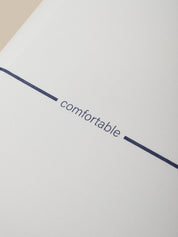 A close-up of a pillow with the word 'comfortable' printed in dark navy blue lowercase text, centered between two short navy blue horizontal lines.