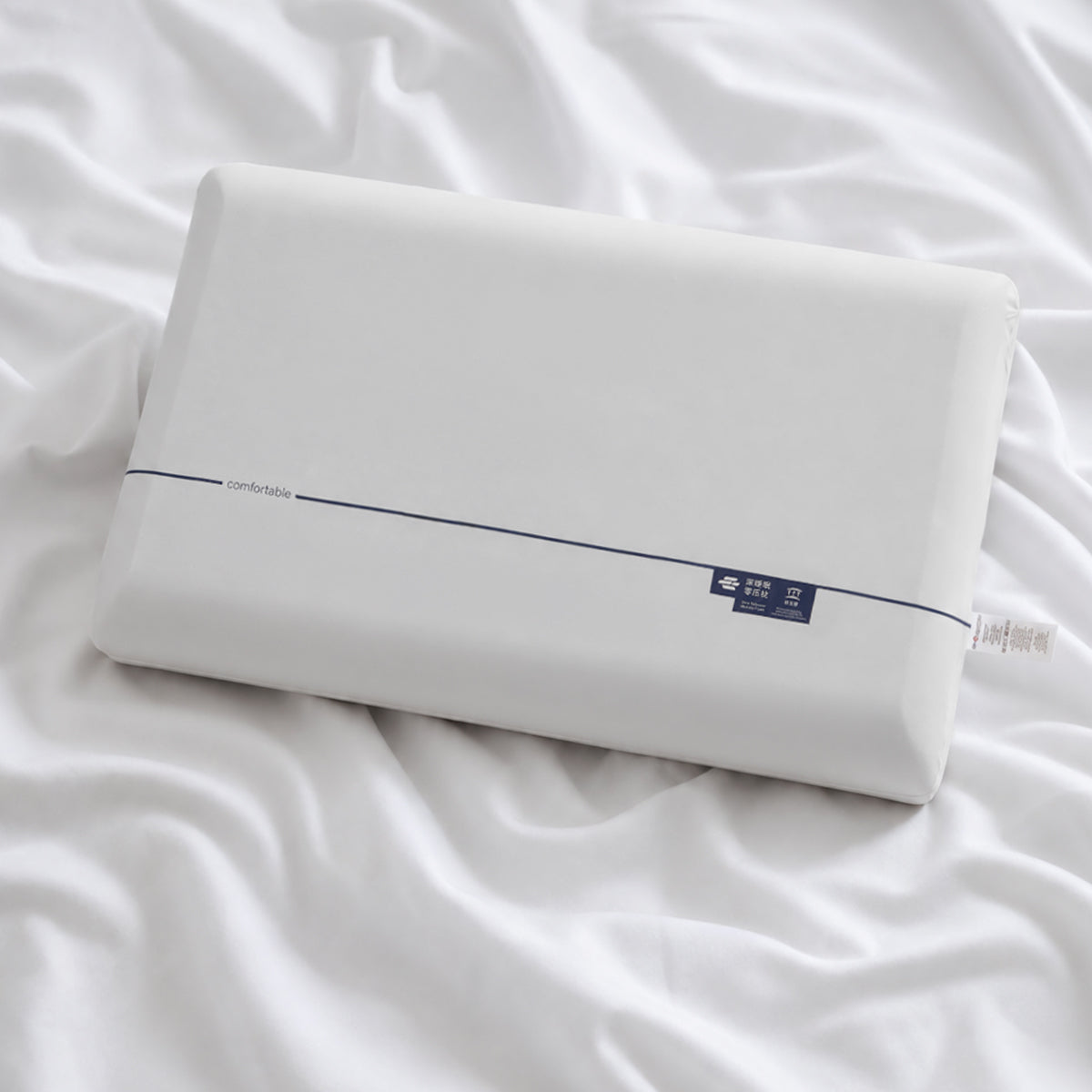 A white rectangular pillow resting on rumpled white bedding, with a thin dark stripe and a small dark blue label near one end.