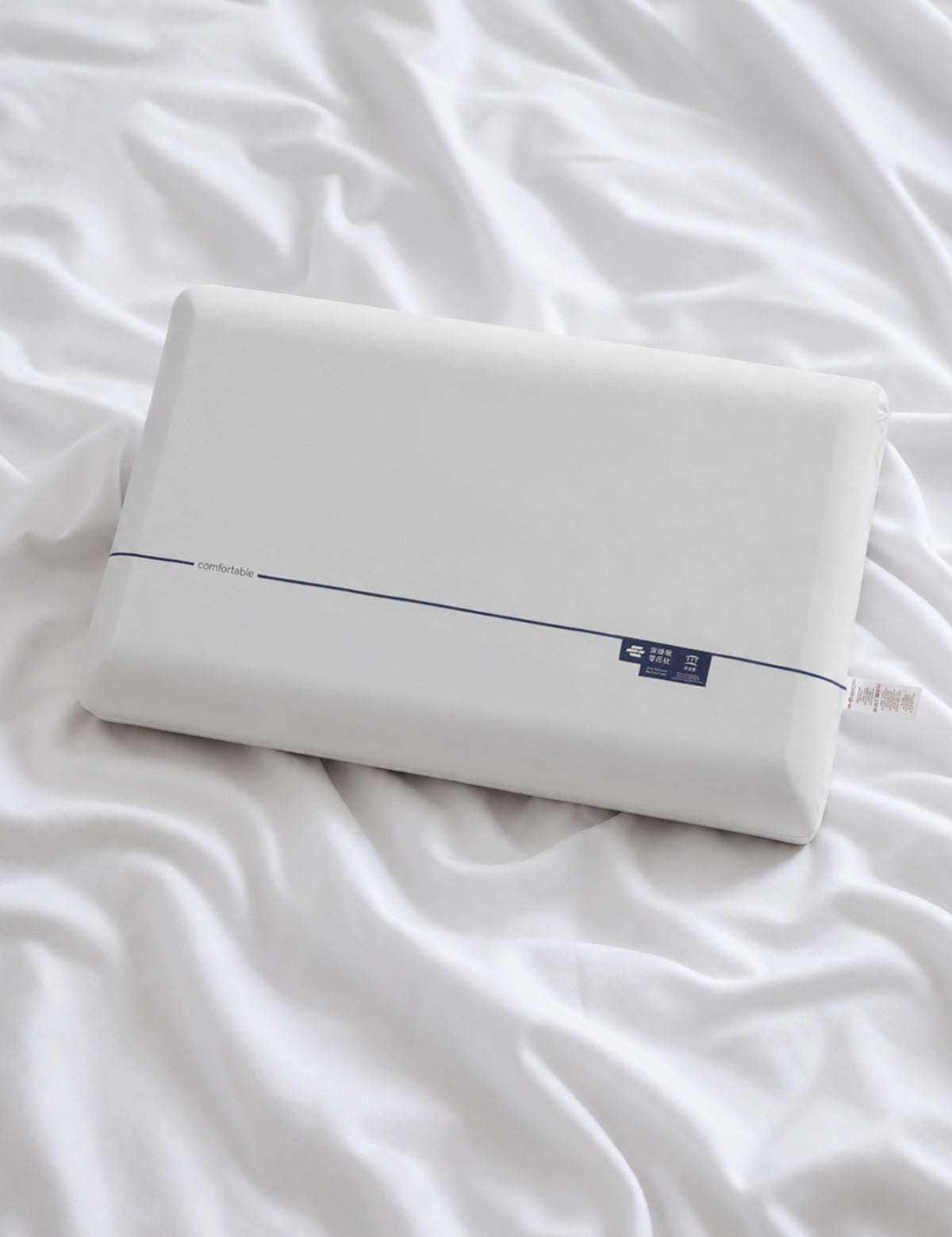 A white rectangular pillow resting on rumpled white bedding, with a thin dark stripe and a small dark blue label near one end.