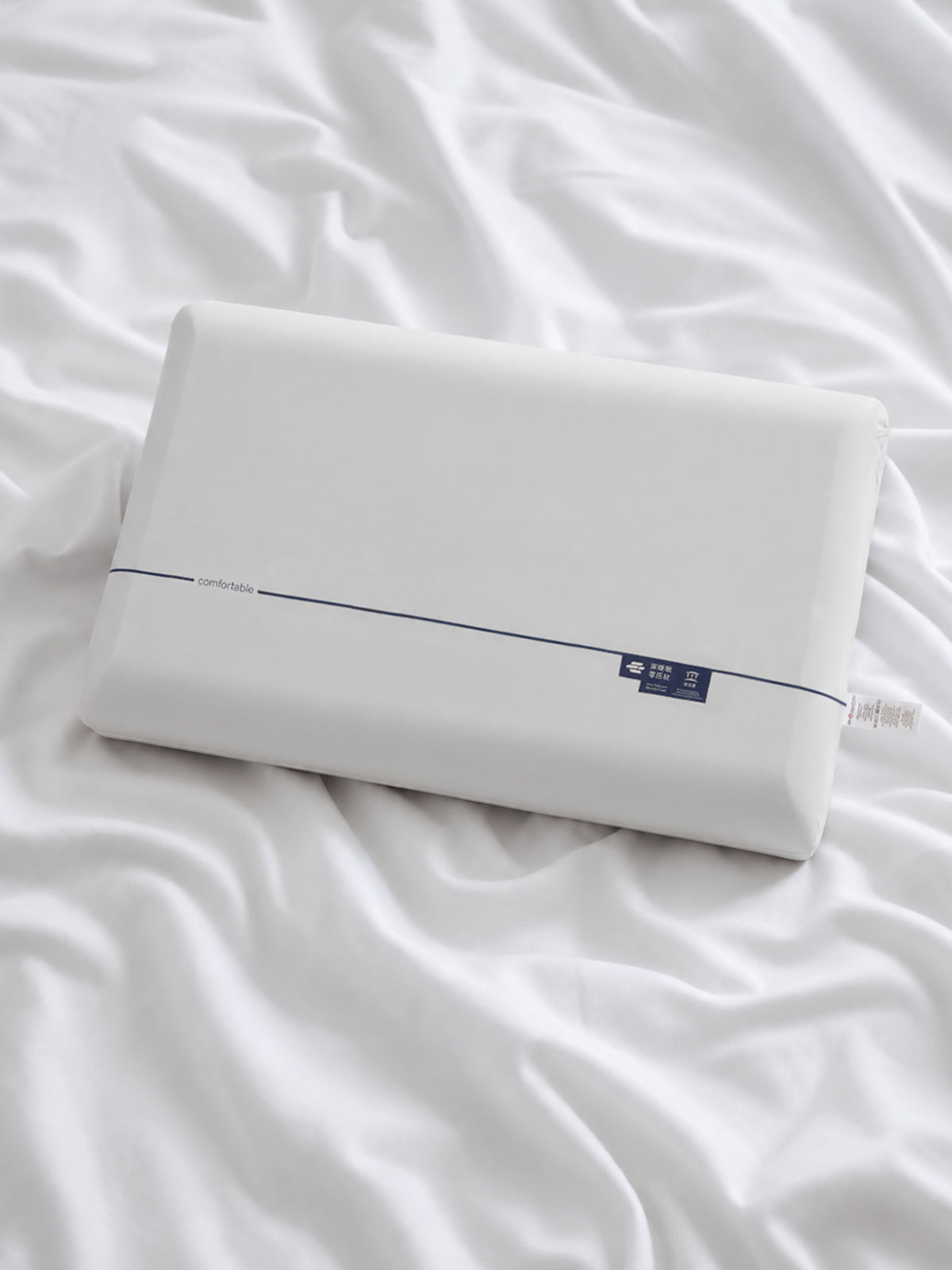 A white rectangular pillow resting on rumpled white bedding, with a thin dark stripe and a small dark blue label near one end.