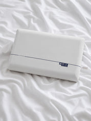 A white rectangular pillow resting on rumpled white bedding, with a thin dark stripe and a small dark blue label near one end.