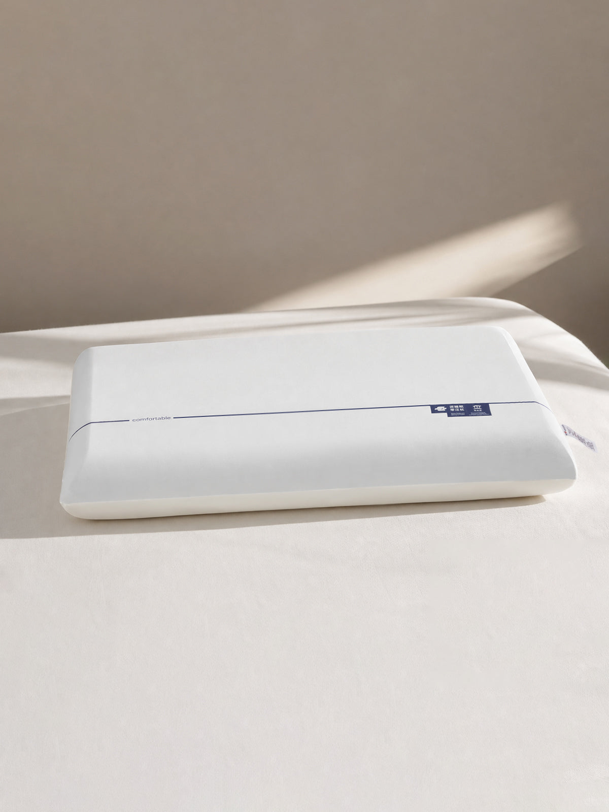 A white rectangular pillow with a small blue brand tag on the right side, resting on a white bed against a beige wall with soft sunlight and shadows.