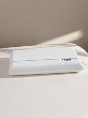 A white rectangular pillow with a small blue brand tag on the right side, resting on a white bed against a beige wall with soft sunlight and shadows.