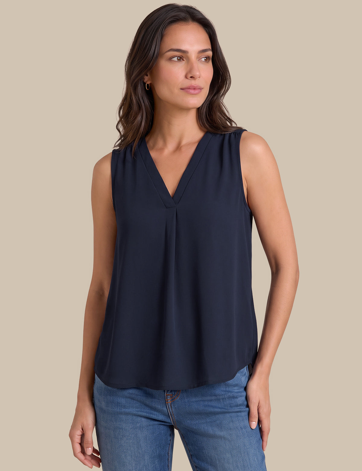 A woman modeling a sleeveless navy blue V-neck blouse with a gathered front, paired with blue jeans, against a solid beige background.