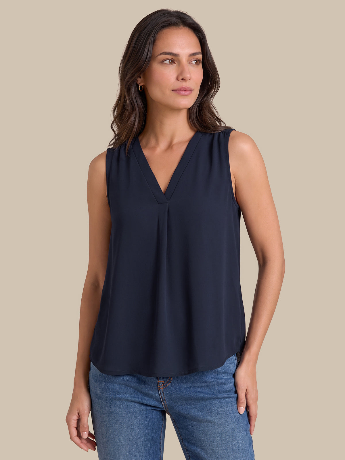 A woman modeling a sleeveless navy blue V-neck blouse with a gathered front, paired with blue jeans, against a solid beige background.