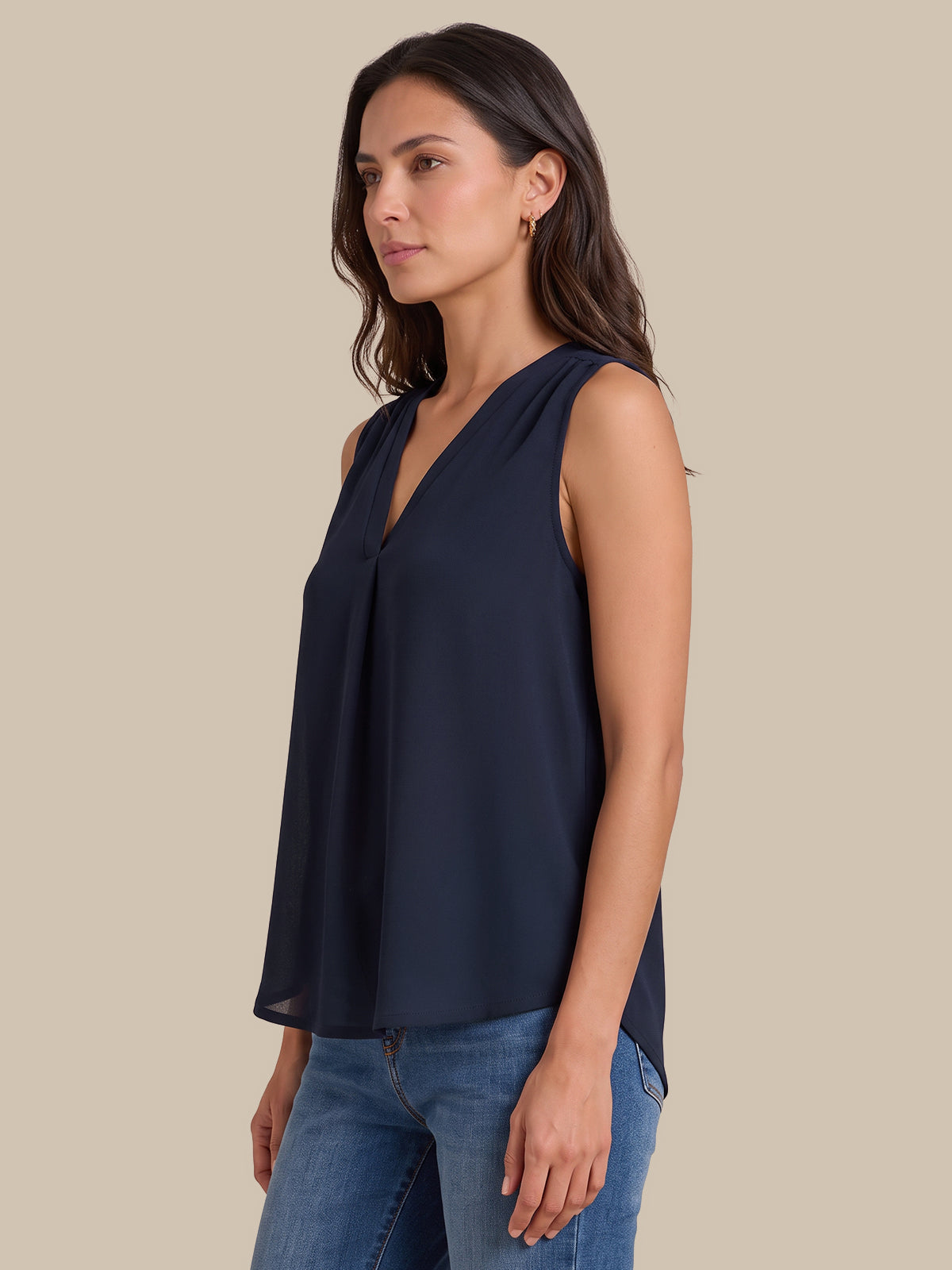 A woman wearing a sleeveless navy blue V-neck blouse with a gathered front, paired with blue jeans, shown against a solid beige background.