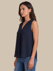 A woman wearing a sleeveless navy blue V-neck blouse with a gathered front, paired with blue jeans, shown against a solid beige background.
