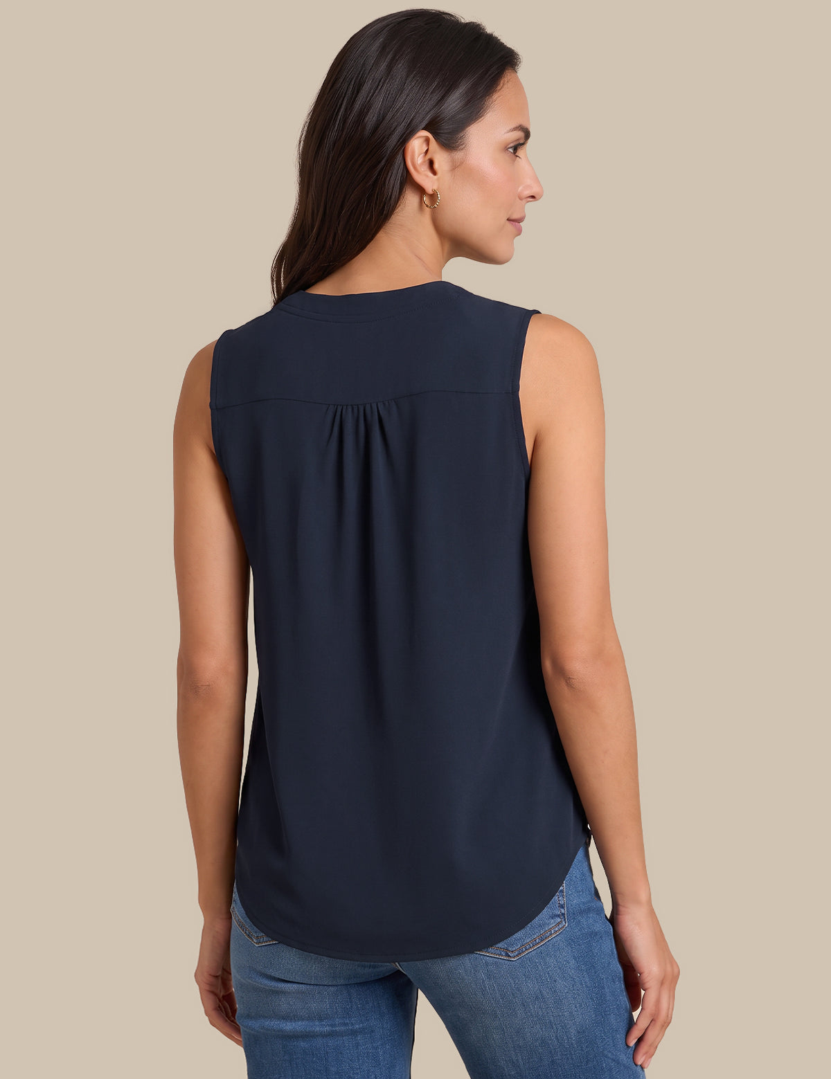 A woman shown from the back wearing a sleeveless navy blue blouse with a gathered back detail, paired with blue jeans, against a solid beige background.