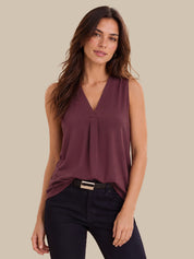 A woman modeling a sleeveless V-neck top in a muted plum purple color, paired with dark pants, against a solid beige background.