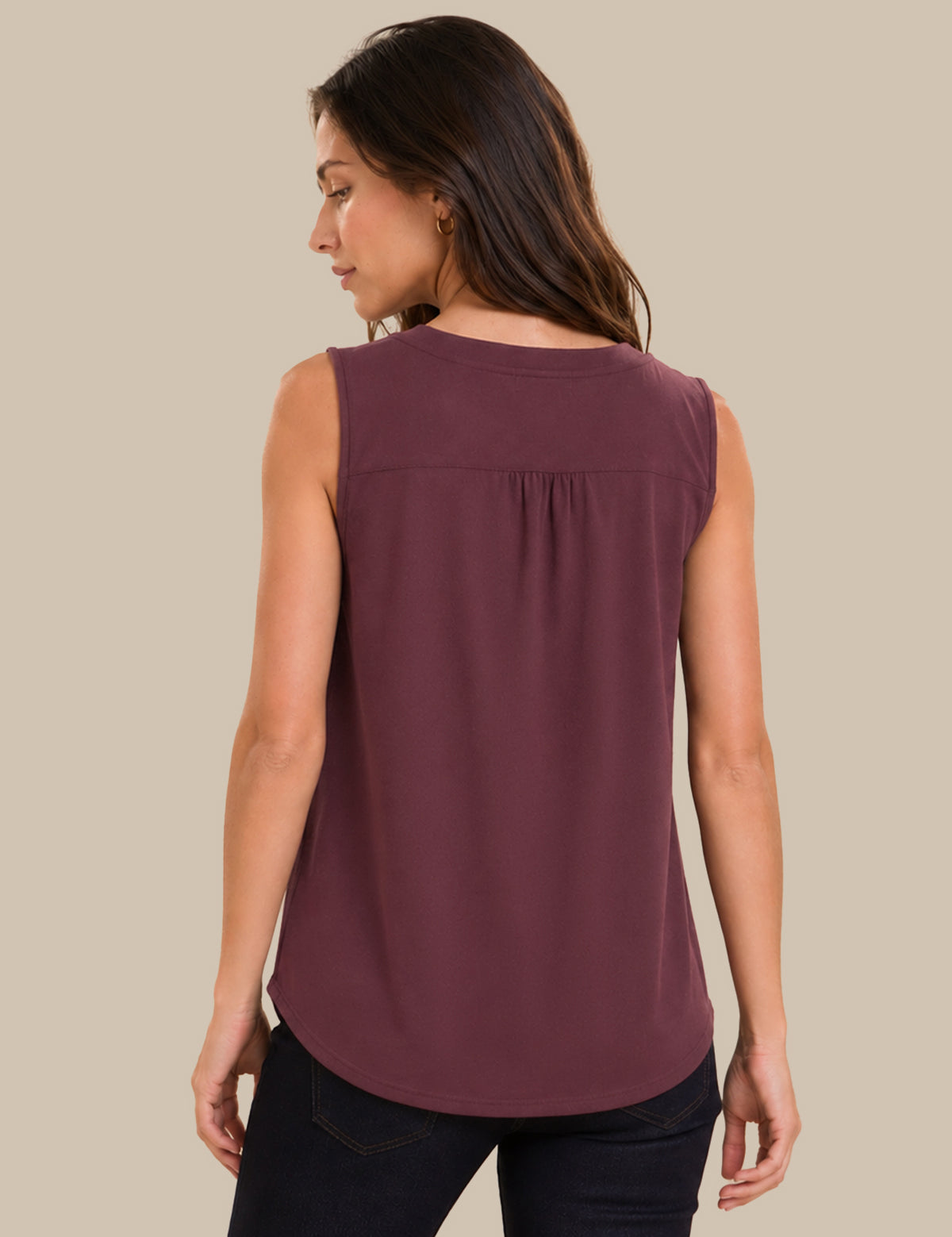 A woman shown from the back wearing a sleeveless, v-neck top in a dusty plum purple color, paired with dark jeans, against a solid beige background.
