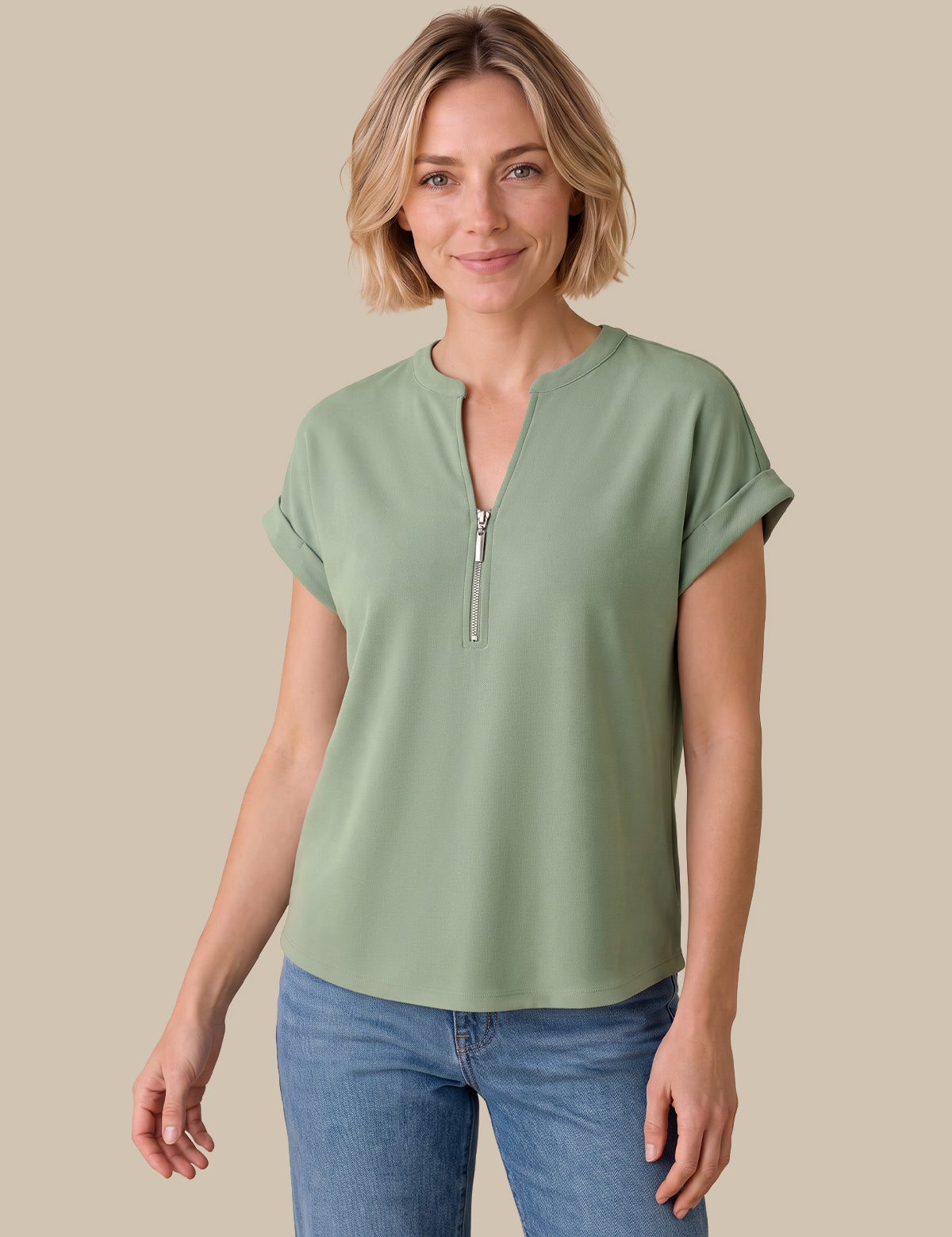 A woman modeling a sage green short-sleeve top with a half-zip neckline, paired with blue jeans, against a solid beige background.