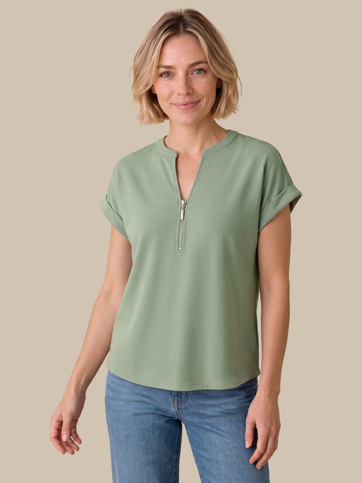 A woman modeling a sage green short-sleeve top with a half-zip neckline, paired with blue jeans, against a solid beige background.