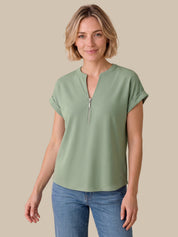 A woman modeling a sage green short-sleeve top with a half-zip neckline, paired with blue jeans, against a solid beige background.