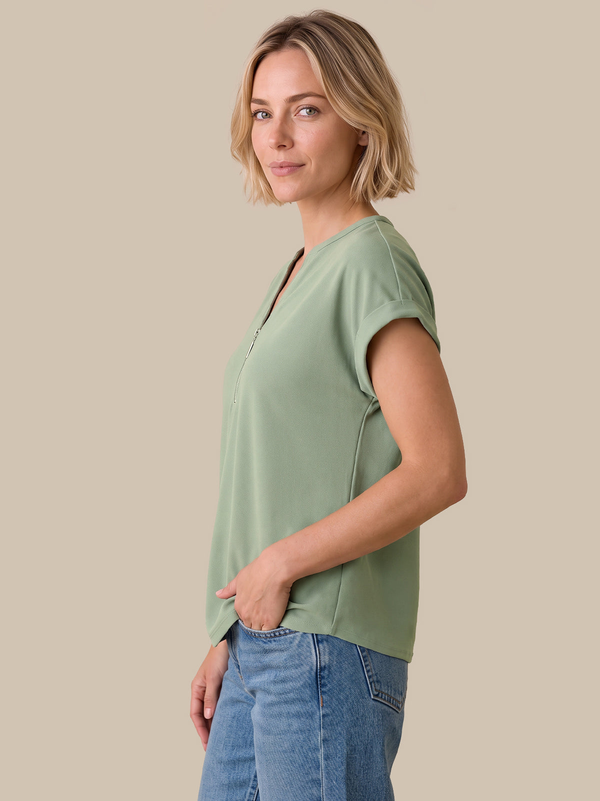 A woman with blonde hair is shown in profile wearing a sage green short-sleeve top and blue jeans, set against a solid beige background.