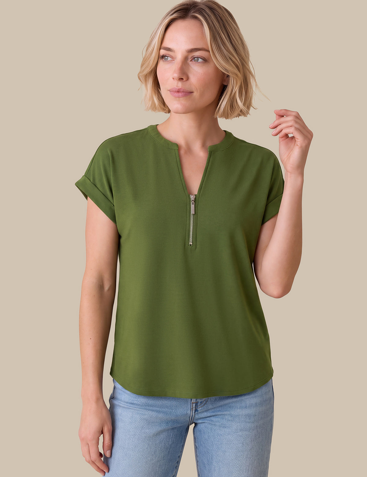A woman modeling a short-sleeve olive green top with a half-zip neckline, paired with light blue jeans, against a solid beige background.