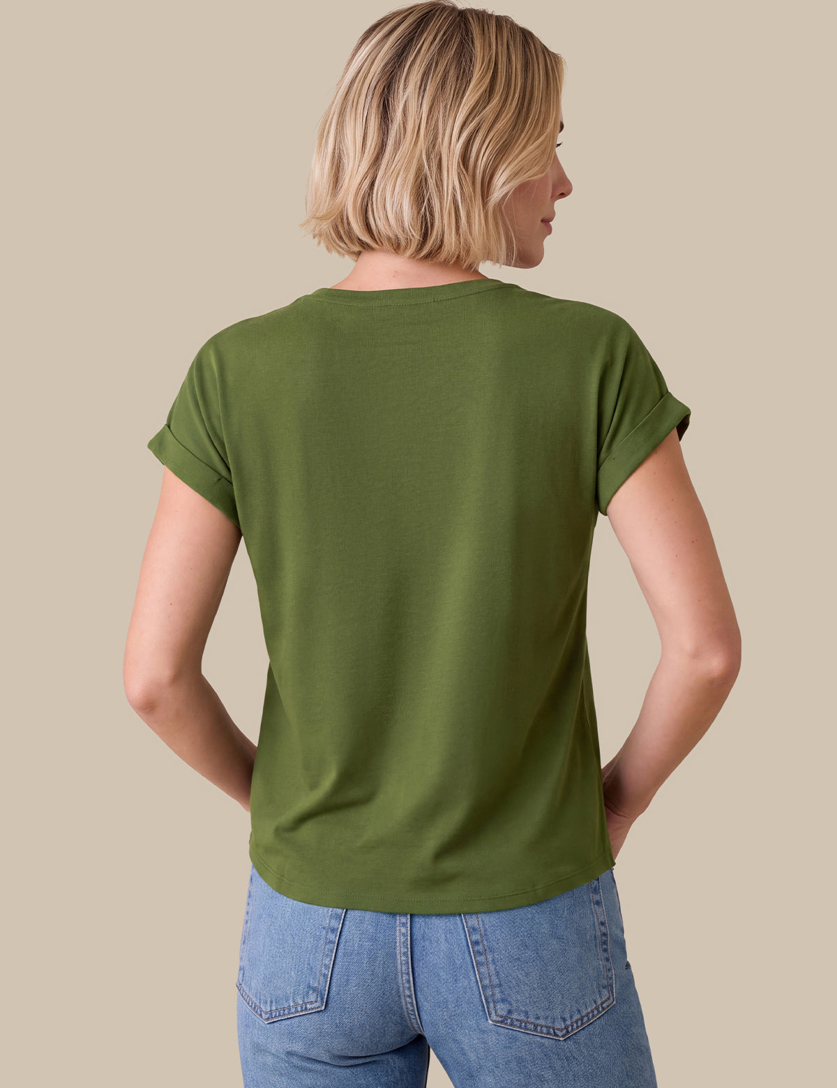 A woman with short blonde hair is shown from the back wearing an olive green short-sleeve t-shirt and blue jeans, set against a solid beige background.