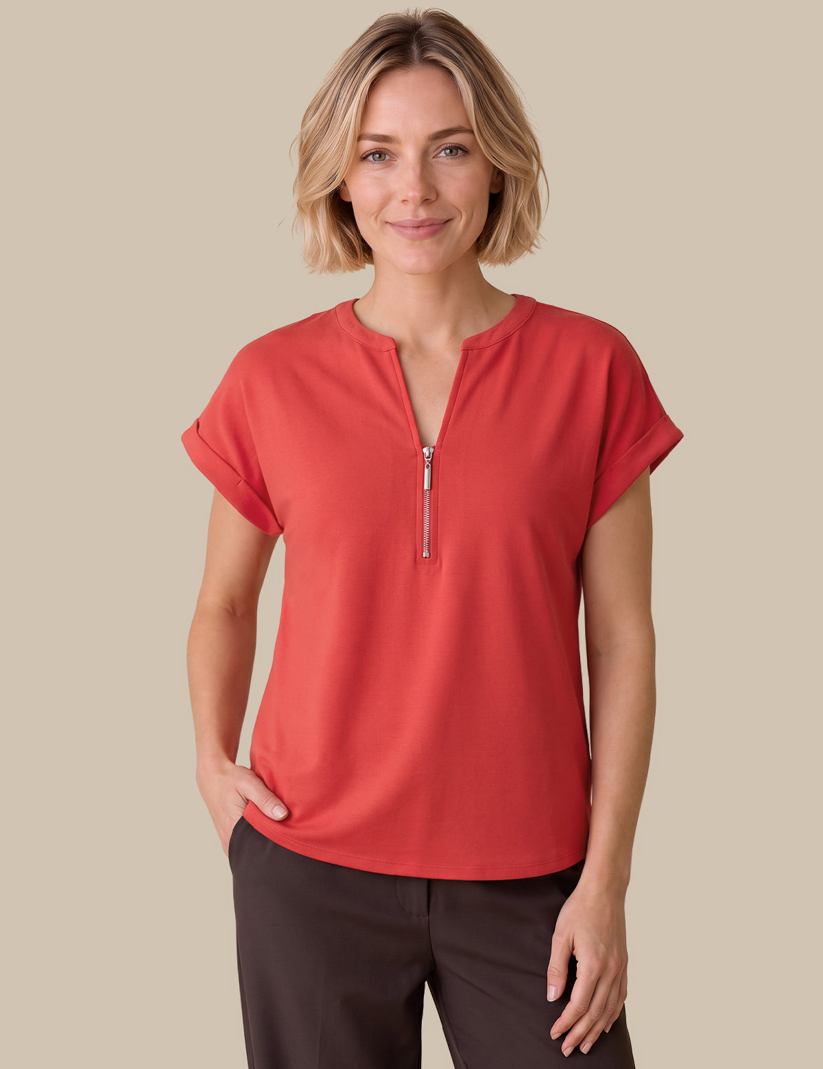 A woman modeling a coral red short-sleeve top with a half-zip neckline, paired with dark brown pants, against a solid beige background.