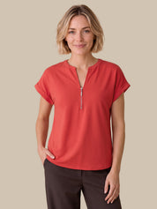 A woman modeling a coral red short-sleeve top with a half-zip neckline, paired with dark brown pants, against a solid beige background.