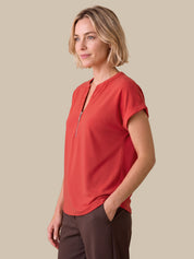 A woman wearing a coral-red short-sleeve top with a half-zip V-neck and dark brown pants, standing against a solid beige background.