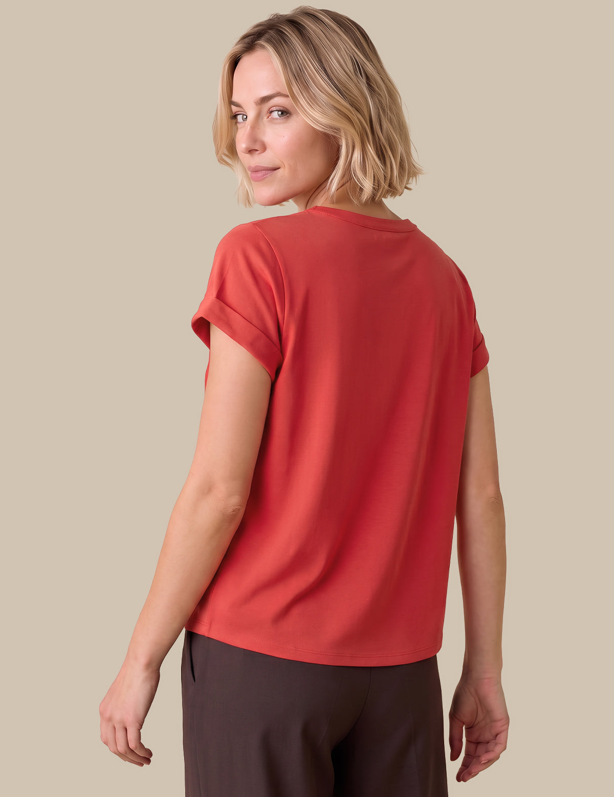 A woman with blonde hair is shown from the back wearing a short-sleeve coral-red top and dark brown pants, set against a solid beige background.