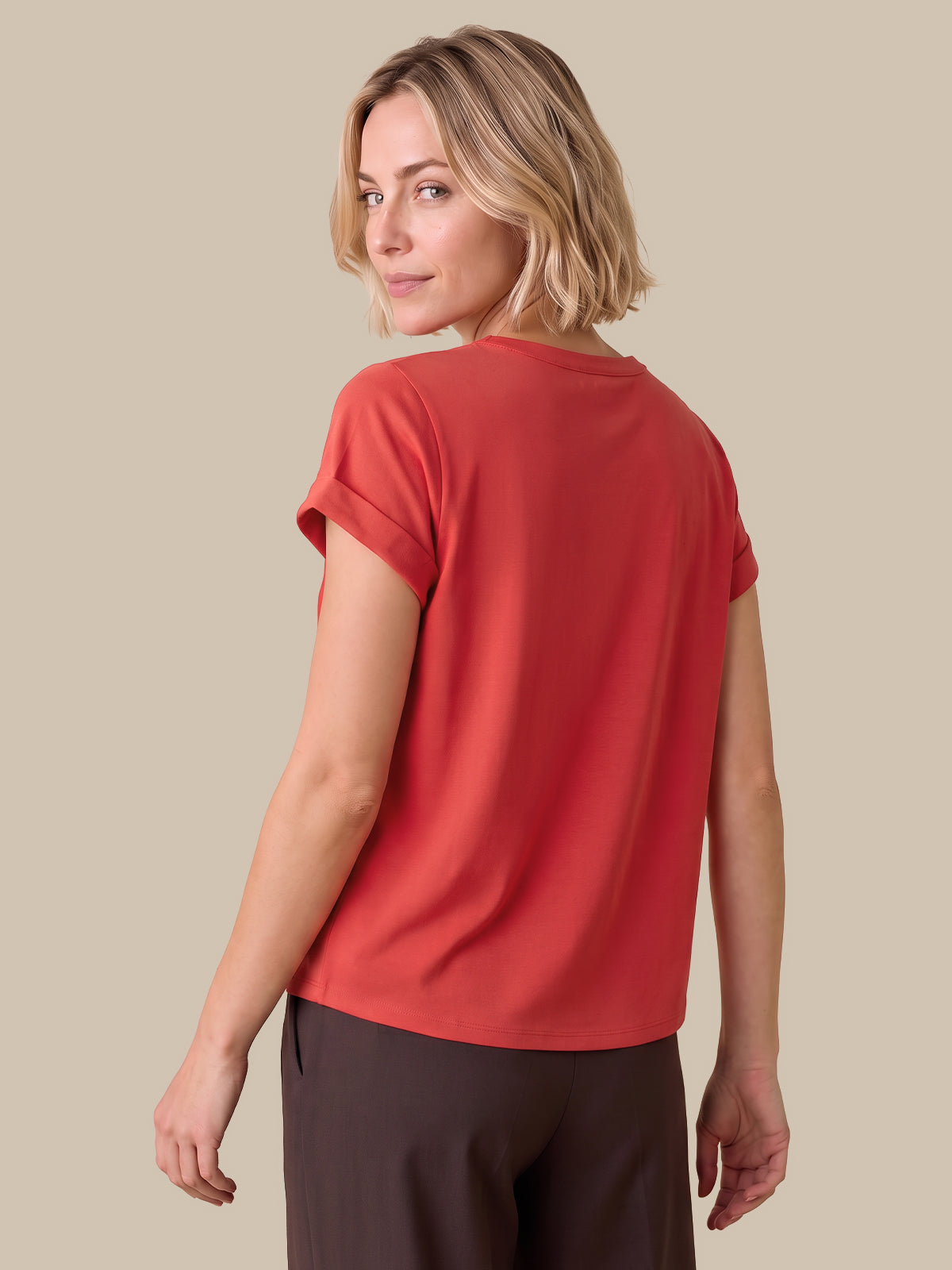 A woman with blonde hair is shown from the back wearing a short-sleeve coral-red top and dark brown pants, set against a solid beige background.