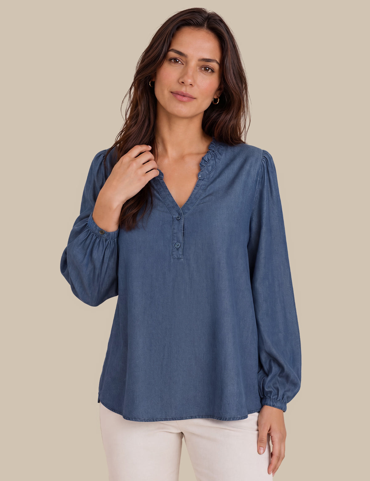 A woman modeling a dusty blue long-sleeve blouse with a ruffled V-neck and button placket, paired with off-white pants, against a solid beige background.
