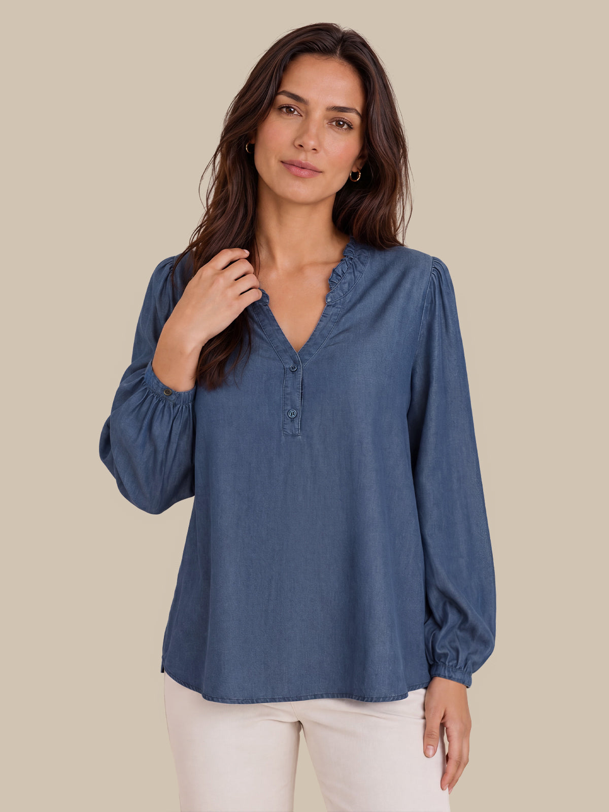 A woman modeling a dusty blue long-sleeve blouse with a ruffled V-neck and button placket, paired with off-white pants, against a solid beige background.