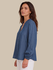 A woman wearing a blue long-sleeve blouse with a split neckline and ruffled trim, paired with white pants, photographed against a solid beige background.