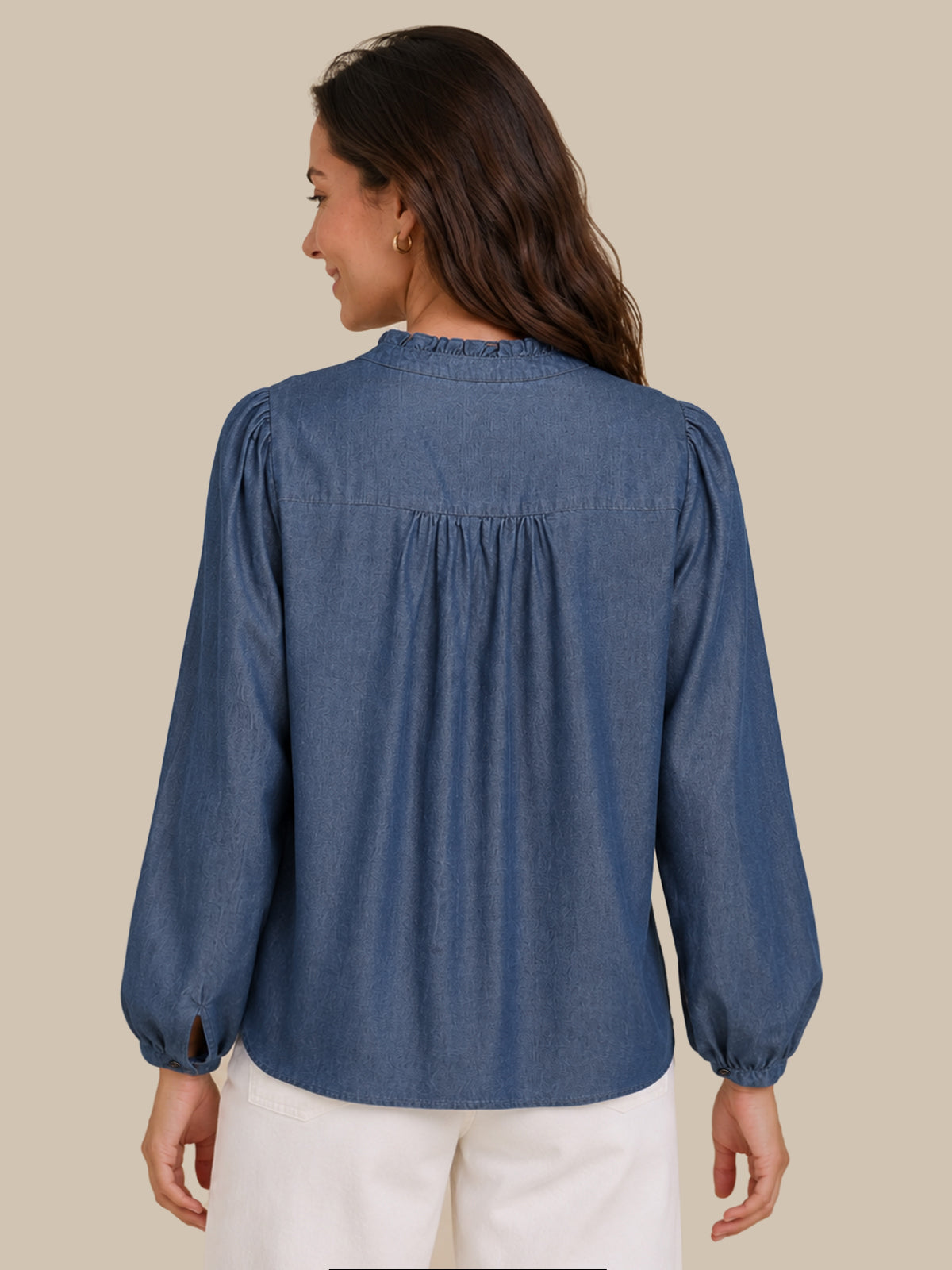 A woman shown from the back wearing a dusty blue long-sleeve blouse with a gathered, pleated back and white pants, set against a solid beige background.