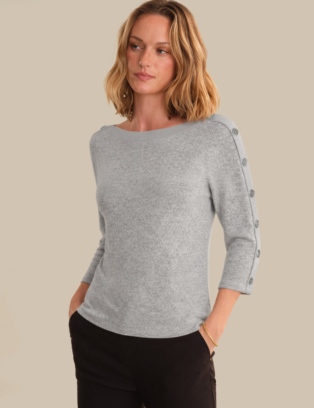 A woman wearing a light gray heathered knit sweater with three-quarter sleeves and decorative buttons down one sleeve, paired with black pants, against a solid beige background.
