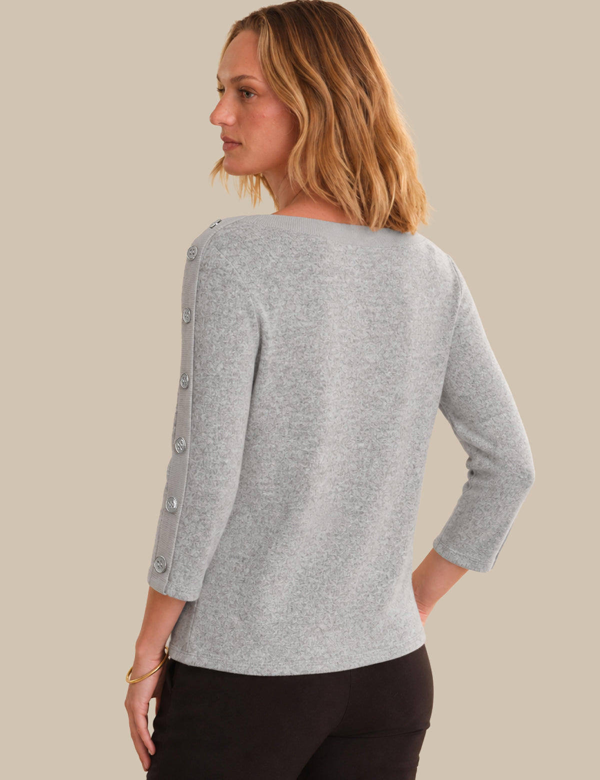 A woman shown from the back wearing a light heather gray knit sweater with decorative silver-tone buttons down the sleeves, paired with black pants, against a solid beige background.