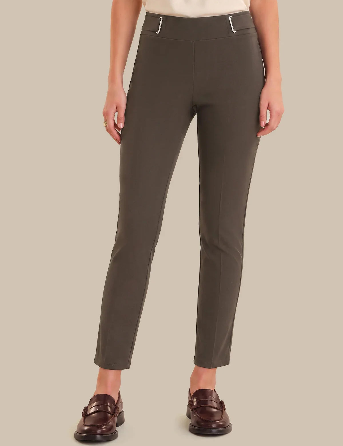 A model wearing slim-fit, ankle-length trousers in a dark olive brown color, styled with a cream top and dark brown leather loafers against a solid beige background.
