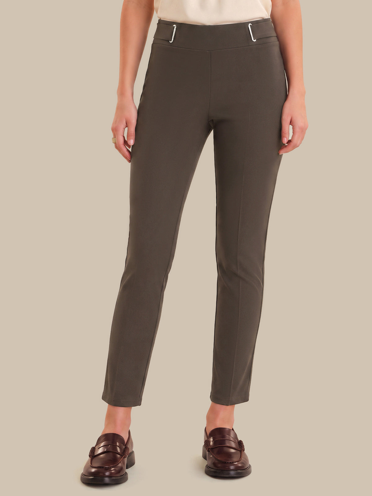 A model wearing slim-fit, ankle-length trousers in a dark olive brown color, styled with a cream top and dark brown leather loafers against a solid beige background.