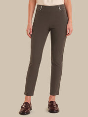 A model wearing slim-fit, ankle-length trousers in a dark olive brown color, styled with a cream top and dark brown leather loafers against a solid beige background.