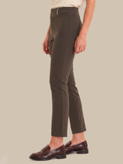 A model is shown from the waist down wearing slim-fit, ankle-length trousers in a dark taupe-brown color, paired with dark brown leather loafers against a solid beige background.