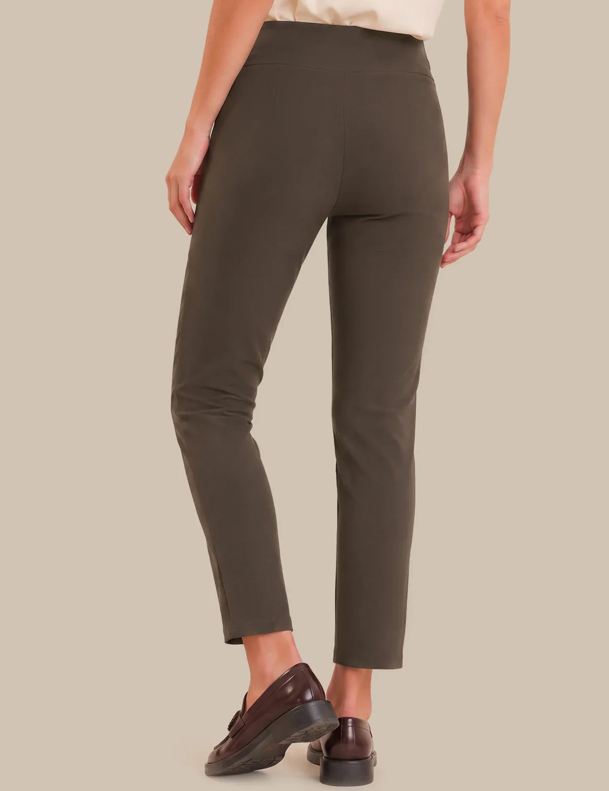 A person shown from the back wearing slim-fit cropped trousers in a dark olive brown color, paired with dark brown leather loafers, against a solid beige background.