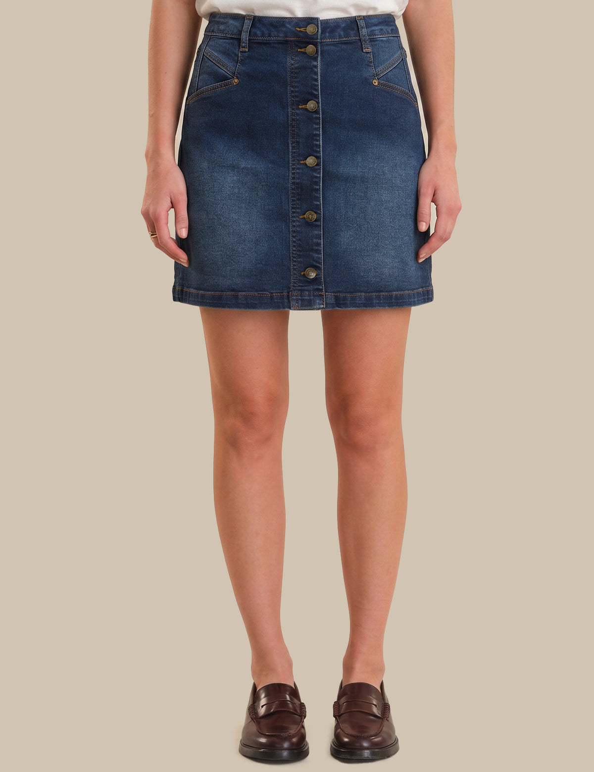 A model wearing a dark blue denim button-front mini skirt, styled with a white top and dark brown leather loafers, set against a solid beige background.